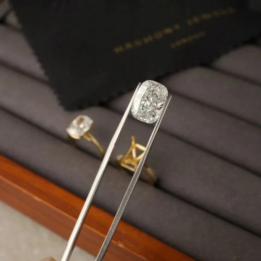 Cushion cut diamond held in tweezers above two yellow gold engagement ring settings
